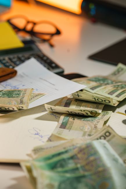 A cluttered office desk surface with scattered £100 banknotes, unorganized papers, a black calculator, and a black mouse pad. The surface appears to be a light-colored wooden or laminate table, with 