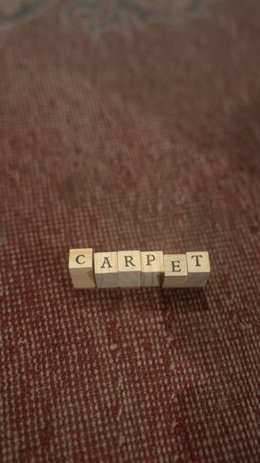 A close-up view of a textured red carpet in an indoor setting, with the surface appearing clean and well-maintained. On the carpet, there are small wooden blocks spelling out the word 'CARPET,' with e