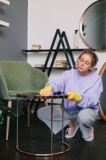 Close-up of a person using a handheld vacuum cleaner to thoroughly clean a green upholstered armchair with a textured fabric surface, situated in a living room with a dark gray sofa in the background.